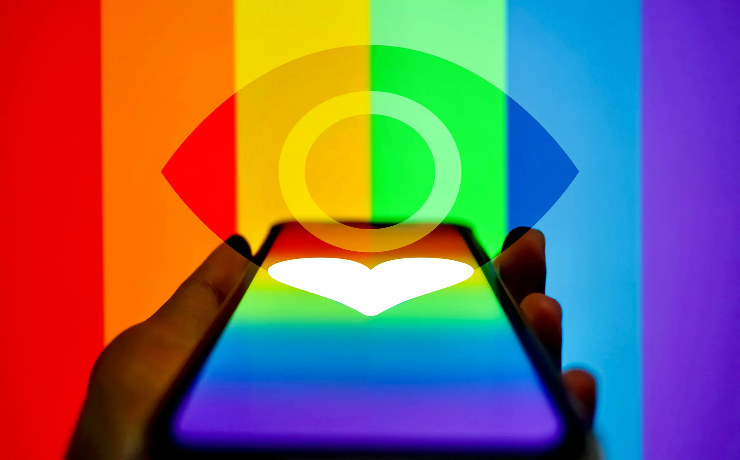 Photo of a hand holding a phone with a rainbow gradient and a white heart on it. The hand holds the phone in front of a vertical rainbow background with an open eye displayed transparently over it, symbolizing surveillance.