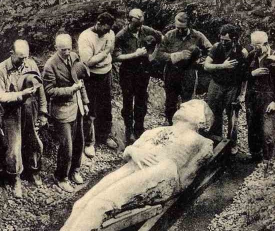 men surrounding a stone sculpture of a man in a hole in the ground doffing their hats